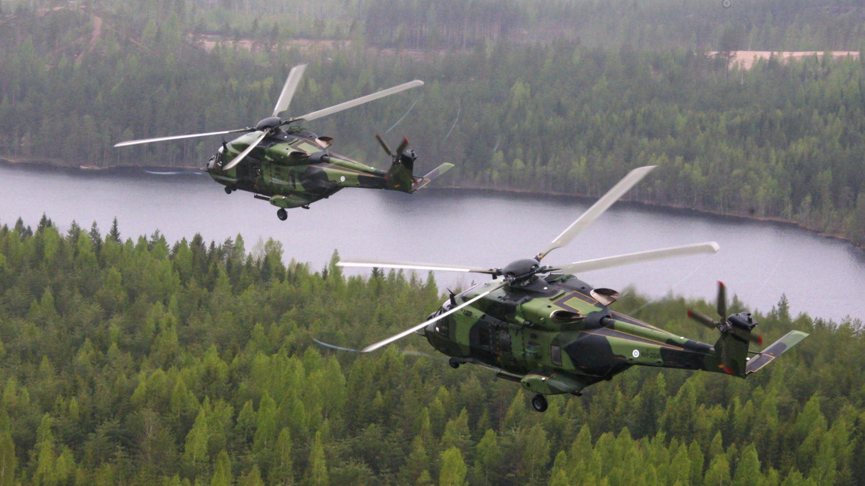 Kuvassa kaksi NH90-helikopteria lentää metsän ja vesialueen yli.
